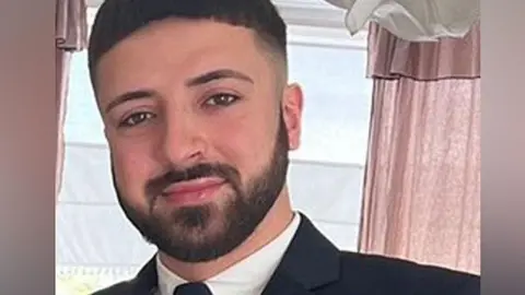 Hertfordshire Police Head and shoulders photo of Kyle Clifford smiling at the camera wearing a black jacket and tie and white collared shirt.