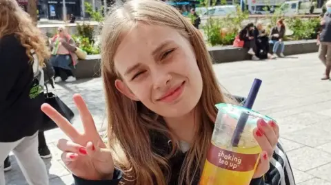 Family handout Image of Rhianan. She is holding up a bubble tea and is smiling with a peace sign for the camera.