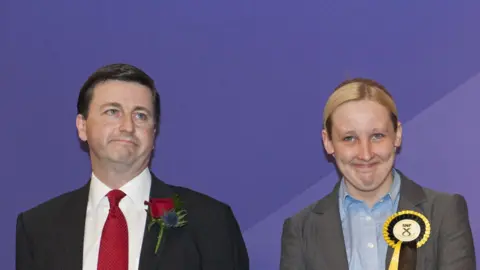 Getty Images The election count in 2015 - Douglas Alexander - a dark haired man with a suit, red tie and red rosette, stands next to Mhairi Black, who has blonde hair and is wearing a grey suit and an SNP rosette