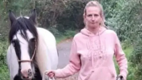 South Wales Police Leanne Williams wearing a pink hoodie. She is holding a rope guiding a brown and white horse beside her. They are in a wooded area.
