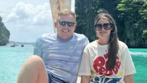 Supplied Tommy Hunter and Chloe Mefford sitting, in shades, by a clear blue sea