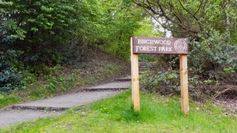 David Dixon/Geograph A path slopes upwards into a wooded area, marked by a wooden sign with 'Birchwood Forest Park' carved into it. 