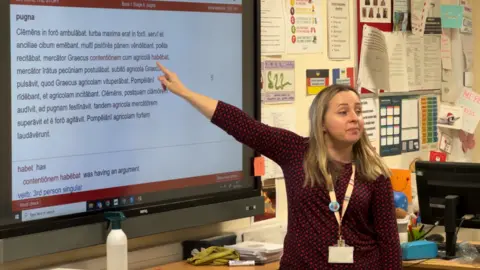 BBC Teacher Marcella Jackson in a classroom. She is pointing behind her to a large TV screen containing Latin words. She has long blonde hair and wears a three-quarter length black top covered in a red spotted pattern and is looking out at the class. She wears a lanyard around her neck.