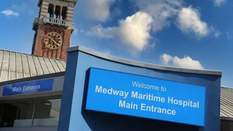 Medway Maritime Hospital A building entrance with a clocktower. A blue and white sign reads "Welcome to Medway Maritime Hospital Main Entrance"