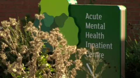 BBC Sign that read "Acute Mental Health Impatient Centre" behind some bushes in front of a red brick wall.