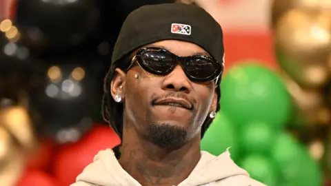 Getty Images Offset in December 2024. He wears a black cap twisted back, dark sunglasses, and a zip-up beige hoody. He has a tattooed neck, a cropped beard and diamond earrings. He's pictured in front of a display, made up of black, green, gold and red balloons.