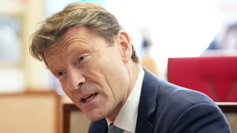 BBC Richard Tice, who has blonde hair, wearing a dark jacket with a white shirt and green tie