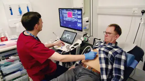 BBC Phil Brown, who has brown hair and glasses, lying down wearing grey jeans and a blue checked shirt that is open at the waist. A technician wearing red scrubs and blue gloves holds the ultrasound device to Phil's stomach while he examines the results on screen