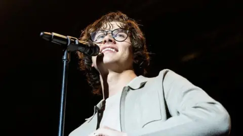 Getty Images Singer David Kushner wearing black rimmed glasses and a grey shirt with a microphone in front of him, smiling and looking out into the crowd 
