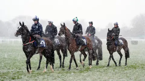 PA Media Horses striding in the snow on Cheltenham Racecourse, with jockeys sitting on top