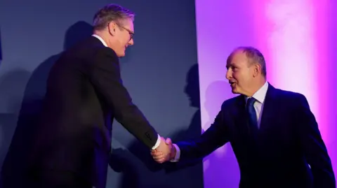 Reuters Micheál Martin said Sir Keir Starmer smiling and shaking hands in front of a blue backdrop with purple lighting. They are both wearing suits.