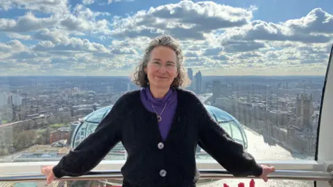 BBC Julia Barfield, a woman with short, wavy grey hair stands inside a capsule on the London Eye; in the background the bright sky is full with clouds and there is a view of the Houses of Parliament