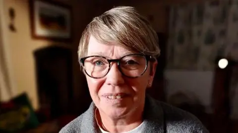 BBC Amanda McKay, in her late 50s, with glasses and a grey top, with a dark background  