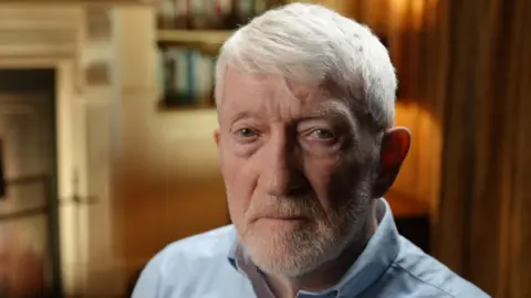 BBC Brendan Hughes looks directly into the camera. He has grey hair and a short beard with a mix of green and brown in his eyes. Brendan is wearing a blue shirt with the top button open. Behind him is an out of focus room with long curtains, a fireplace and a bookshelf with warm lighting which gives off an orange glow. 