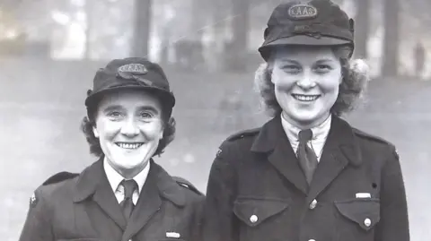 London Ambulance Service Ann Armitage and Betty Leverton looking at the camera. Anne has dark, short hair and wears a cap. Betty is taller and has curled blonde hair and wears a cap.