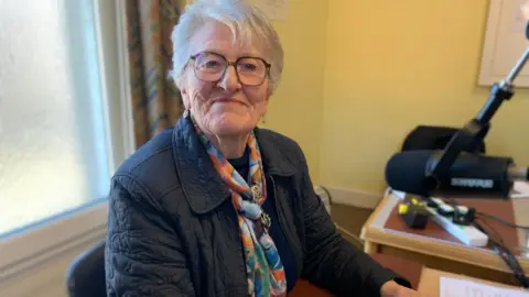 BBC A woman with short grey hair, a navy jacket and a colourful scarf looks towards the camera smiling while sitting in front of a microphone