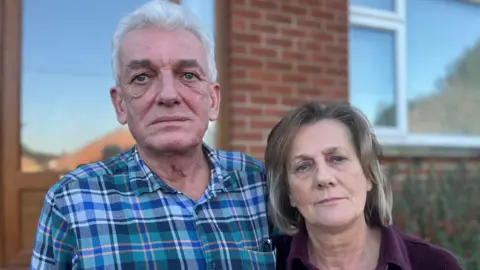 BBC A man and a woman are standing next to each other outside a house. The man is wearing a checked top and sunglasses are in his pocket. The woman is wearing a purple top.