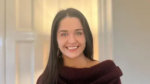 BBC Suzy Spence. She has long dark brown hair, blue eyes and smiling directly into the camera and wearing a dark red jumper.