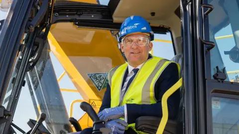 Ministry of Justice James Timpson sitting in the cab of a construction vehicle. He is wearing a blue hard hat and a yellow high-vis jacket over a navy suit. He is wearing clear protective goggles.