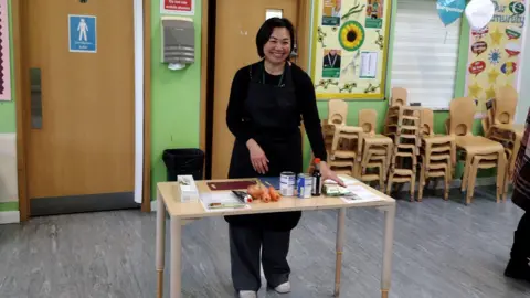 London Borough of Sutton Woman stands at table with ingredients - two carrots, and onion, tomato paste and some cans of food and soy sauce. There are also some recipe sheets on the table. The woman is smiling and is wearing black top and trousers. The room she is standing in has rows of stacked up chairs and there is a door to the men's loos in the background and some posters on the wall.