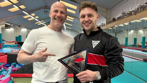 Johhny Reed/BBC Joe Fishburn and Roddy Mackay pose for a photo with the Gladiators trophy. Mr Mackay, who is smiling in the picture, is bald and wearing a white t-shirt.