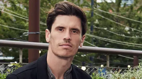 Forest Live A man with dark hair and wearing a black jacket and grey shirt, stands in front of some fencing. A telegraph pole and trees can be seen in the background.