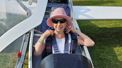 Angel Clark Maureen Clark is pictured sitting in the front seat of a white glider as it rests on a grass field. She smiles at the camera. She wears a pink bucket hat and a white T-shirt. She wears a parachute.