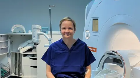 Katy Stephenson Katy Stephenson is pictured in a hospital room wearing a dark blue hospital scrub top. She sits on the bed of a piece of equipment and smiles at the camera. She has short blonde hair.