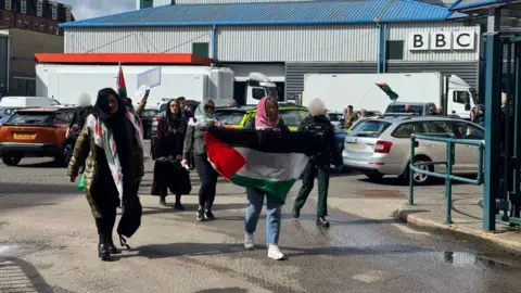 A group of people, with one carrying a Palestine flag, are led out of Blackstaff House by some police officers. The protestors are wearing Keffiyehs over their head. In the background is a large grey building with the BBC logo on it.