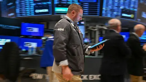 Getty Images A trader walks past holding a tablet on the floor of the New York Stock Exchange (NYSE) at the opening bell on April 21, 2025, in New York City. Behind him are blurred blue screens showing the markets and men in black suits.