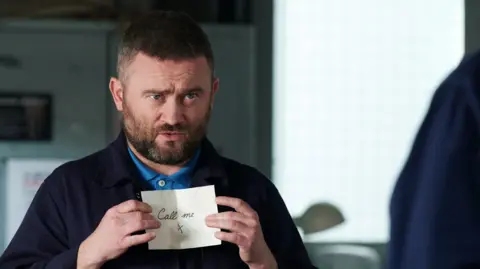 BBC Studios Stephen Purdon as Bob O'Hara in River City - he is holding up a scrap of paper saying call me and showing it to someone out of the camera shot, with a frown on his face