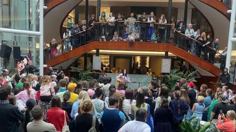A distance shot of Amy MacDonald playing in Glasgow, fans in a crowd in front of her and also on a stairway and landing above her.