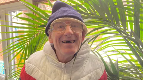 A man wearing glasses and a white puffer jacket, smiling in front of  a large plant.