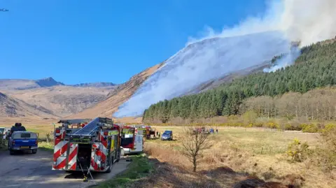 Jethro Lennox Fire engines parked in front of hills that are covered in smoke.