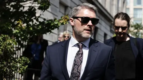 PA Media Adam Smyth, a man with short grey hair, wearing a navy suit, blue shirt and patterned navy and purple tie. He is wearing dark sunglasses, there is a large stone building and green foliage in the background. A dark haired woman, also in a navy blazer, follows behind him.