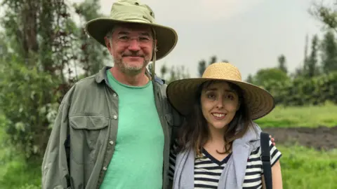 Victoria Gill/BBC News The image shows a man (on the left) and a woman smiling into the camera. Both are scientists working on conservation of the wild axolotl in Xochimilco, close to Mexico City. The background shows the lush, green habitat that is their field site. 