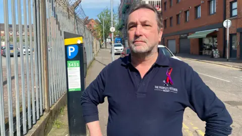 A man with grey longish hair and a beard wears a navy jumper, with The People's Kitchen, Belfast on the lapel. 