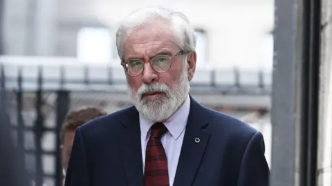 PA Media Gerry Adams, pictured entering a courthouse, he has grey hair and a grey beard. Wearing glasses, a white shirt and navy suit jacket, with a striped patterned red tie.