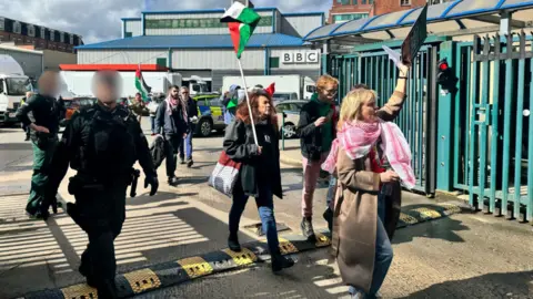 BBC A group of protesters, holding Palestine flags and wearing Keffiyehs, are leaving through large green iron gates accessing a car park near Blackstaff House. There are two police officers on the left of the image, but their faces are blurred.