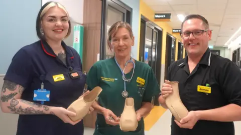 Great Western Hospital NHS Foundation Trust Three healthcare staff members in scrubs in a hospital corridor. They all look at the camera, smiling and all holding a cardboard containers, shaped at the top to allow someone to urinate.