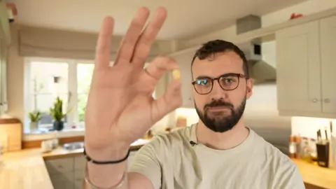 BBC Chris Brookes-Smith has dark brown hair and beard, and holds up a peanut in his kitchen at home, with a window in the background and light grey cupboards on the walls behind him