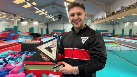 Johnny Reed/BBC Joe Fishburn, who has dark hair and a stocky build. He is holding a triangular golden trophy with the letter G inside the shape.