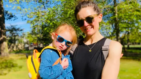 BBC A mother holding her two-year-old daughter in a park in Aberdeen with several trees in the background. The mother is wearing yellow sunglasses, a black vest and a rucksack with her brown hair tied up. The daughter has a bright yellow backpack, a denim dress and blue sunglasses. She has bright red hair.