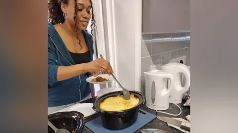 London Borough of Sutton Woman wearing a blue hoodie and black top stirs ingredients in a slow cooker. The slow cooker is black and inside there is what looks liked mashed potato inside. Along the counter you can see two white kettles on the right and on the left a partial view of another slow cooker with a metal serving spoon inside.