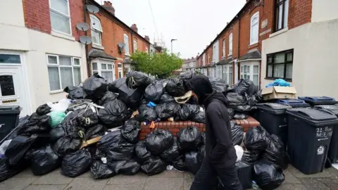 PA Media A man walks past a large pile of black bin bags stacked over a wall on a residential street. There are terraced houses on either side of the road.