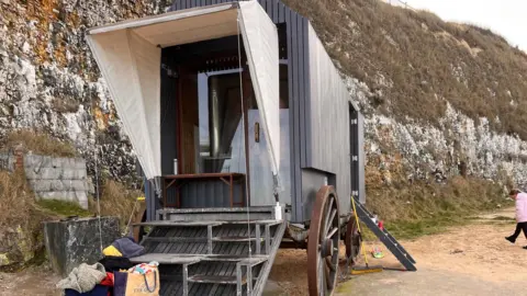 Ian Palmer/BBC An exterior image of a free-to-use sauna in Margate which has attracted visitors from all over the south east. The sauna is elevated above the ground and has four steps up into it. Above the entrance there is a white tarpaulin which extends out. Either side of the sauna are two wheels; two larger wheels at the front and two smaller wheels at the back.