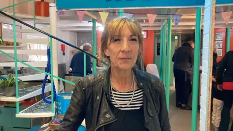 BBC A woman wearing a leather jacket, stood behind a series of green coloured pillars and a set of spiral stairs. 
