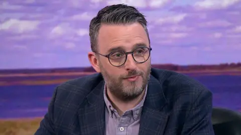 Nick Mathison, with dark hair and beard, wearing a checked blue jacket and purple shirt.