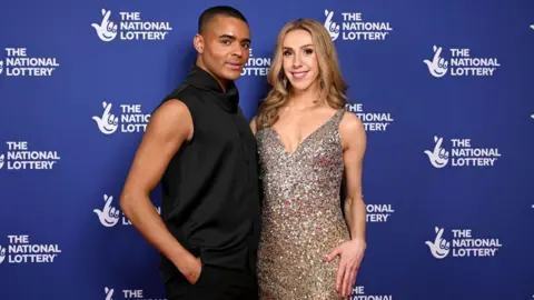 Getty Images Layton Williams and Lauren Drew standing together at the National Lottery's Big Night of Musicals Red Carpet at the AO Arena. Layton is on the left and is wearing a black suit with one arm in his trousers. Lauren is smiling at the camera wearing a sparkly silver dress with curly light brown hair. 