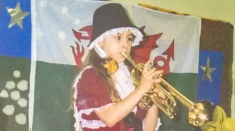 Lauren Drew An old image of Lauren playing the trumpet in a St David's day costume on stage. She is standing in front of a Welsh flag. 